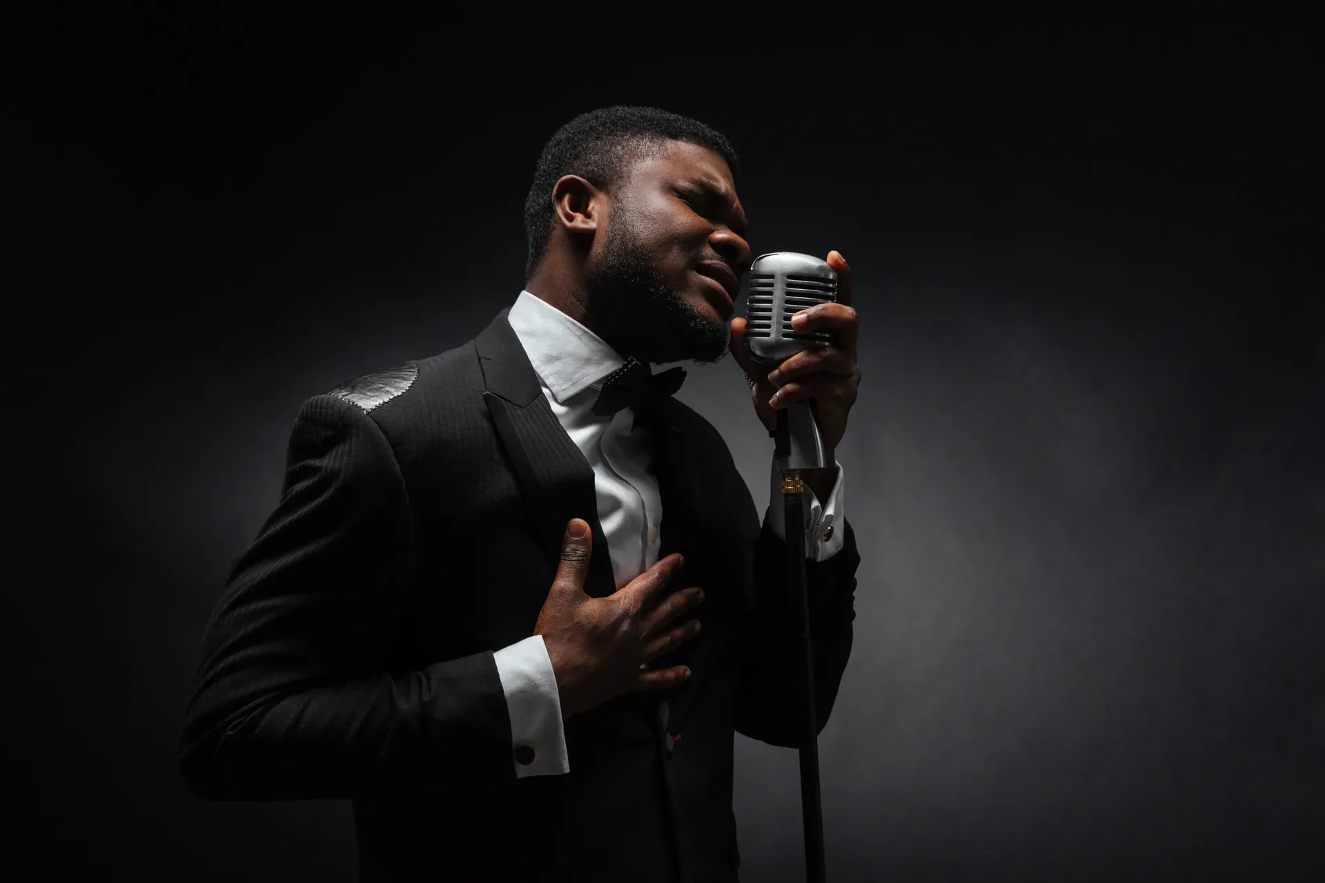A singer in a tuxedo mid-performance at a vintage chrome microphone, hand on chest